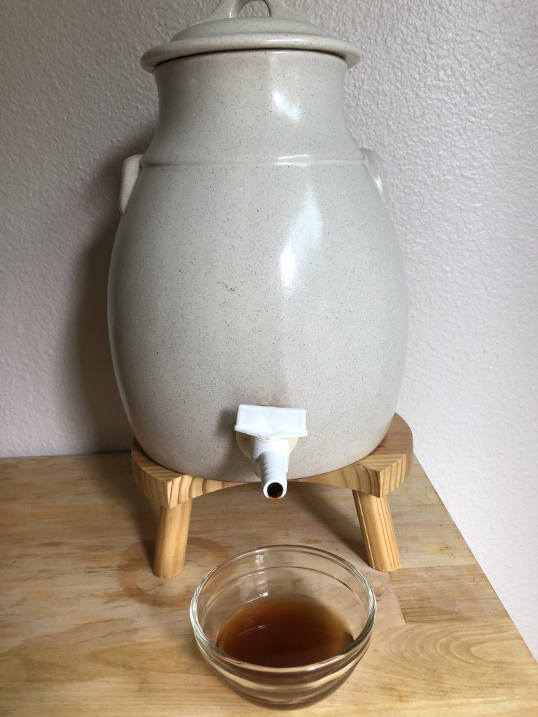 A beige ceramic vinegar crock, with plastic spigot, on top of a wooden stand. Foreground is a small glass bowl with a tiny amount of a brown, hazy liquid - vinegar.