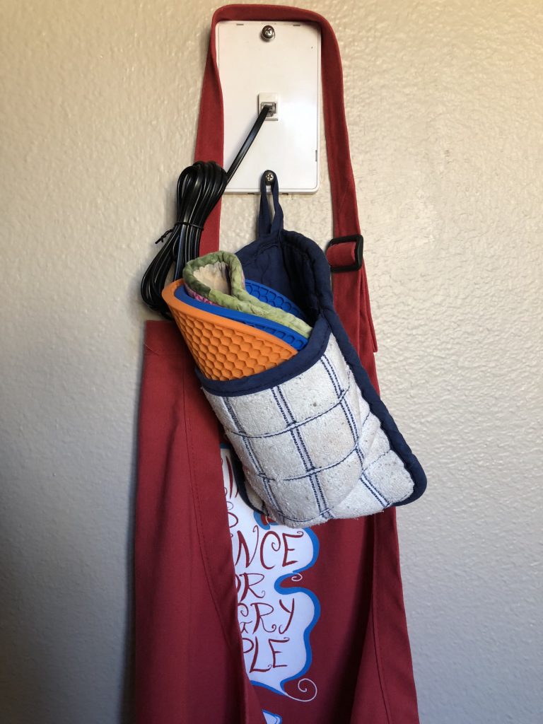 A phone jack holding up a red apron, a black phone cord to nothing, and a potholder with a pouch holding up three other potholders.